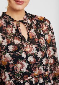 Femme portant un chemisier fleuri avec un détail à noeud, présentant des fleurs blanches, roses et orange sur un fond sombre.