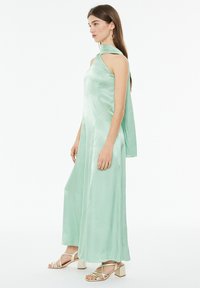Abito in satin verde menta con design a spalla unica, schiena drappeggiata e silhouette fluida. Abbinato a sandali con tacco a blocco e fasce.