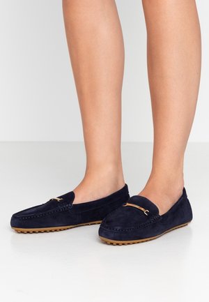 Jambes portant des mocassins en daim bleu marine avec des semelles en caoutchouc beige cloutées et un détail en métal doré sur le dessus, debout sur un sol blanc.