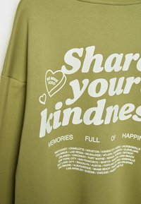 Sweatshirt vert olive avec texte blanc : "Partagez votre gentillesse" et un cœur avec "Sois à moi XOXO," ainsi que des noms de villes en dessous.