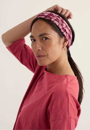 Tumejuustega naine, kes kannab roosat lillemustriga peapaela, kuldseid rõngaskõrvarõngaid ja lahtist roosat toppi, tõstab ühe käe pea taha.