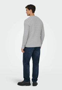 Homme de dos portant un pull à mailles en câble gris clair, un jean bleu foncé et des chaussures noires, sur un fond clair uni.