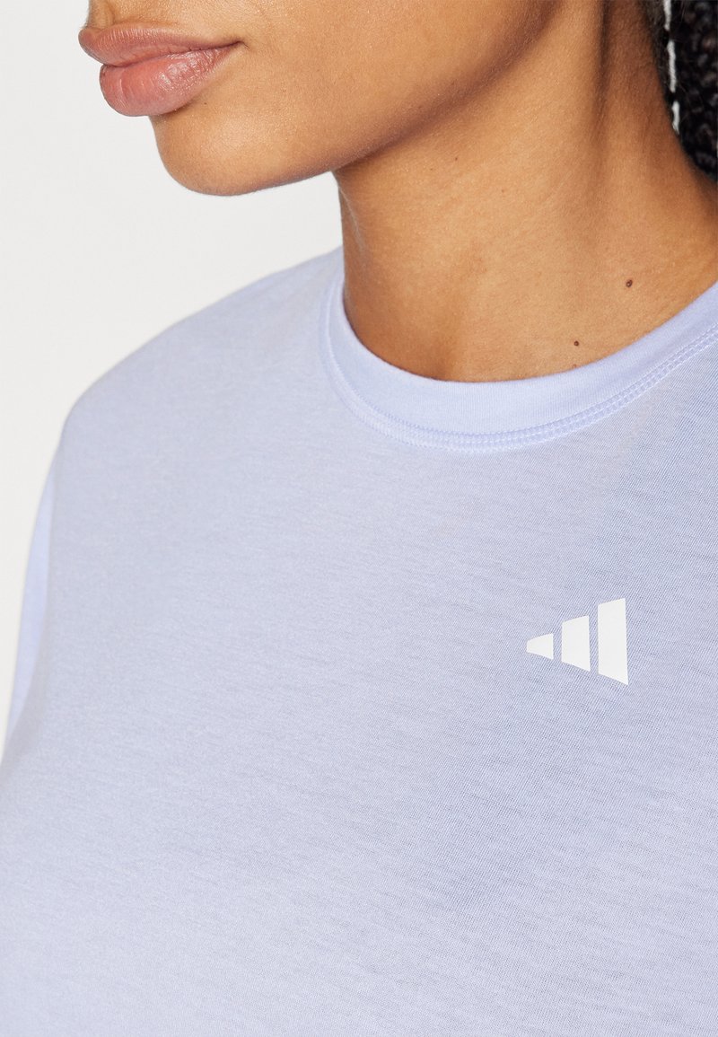 Camisa corta de manga en color púrpura claro con un cuello redondo, que presenta un logo triangular blanco en el área del pecho izquierdo. Textura suave.