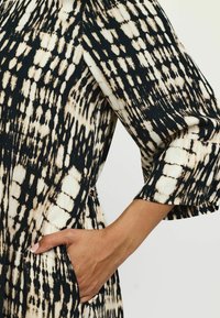 Mustvalge kleit mustri kujundusega, millel on kolmeveerandi varrukad ja küljetasku. Kanga peal on pehme, struktureeritud välimus.