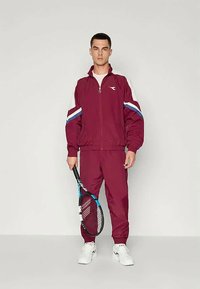 Bordó tréningruha cipzáros kabáttal és összeillő nadrággal, kék és fehér csíkokkal a vállán. Teniszütőt tart a kezében.