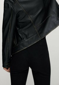 Veste en cuir noire avec une coupe ajustée, présentant des coutures visibles et un design légèrement court. Portée sur un pantalon noir ajusté.