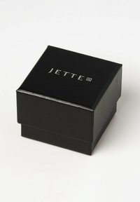Scatola per gioielli in finta pelle nera con texture in rilievo, con logo argentato "JETTE" sulla parte superiore. Forma quadrata, coperchio con cerniera.