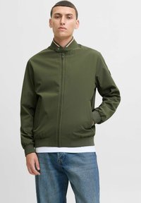 Veste vert olive avec col montant, fermeture éclair à l'avant et poignets élastiques. Tissu texturé et poches latérales, portée sur une chemise blanche.
