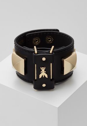 Musta leveä nahkarannekoru, jossa kullattu hyönteisaiheinen merkki ja kultaiset pyramidihelmikoristeet, kiinnitetty kahdella painonapilla, esillä valkoisella alustalla.