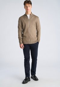 Jeune homme debout portant un pull beige à fermeture éclair, un sous-pull blanc, un pantalon foncé et des mocassins noirs à glands, sur un fond uni.