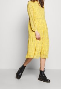Vestido floral amarillo con mangas largas, cintura fruncida, dobladillo en capas, combinado con botas de combate negras con cordones. Presenta un escote tipo ojal.