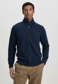 Marineblaues Zip-Up Sweatshirt aus weichem, strukturiertem Material mit hohem Kragen und gerippten Bündchen, kombiniert mit hellbraunen Hosen.