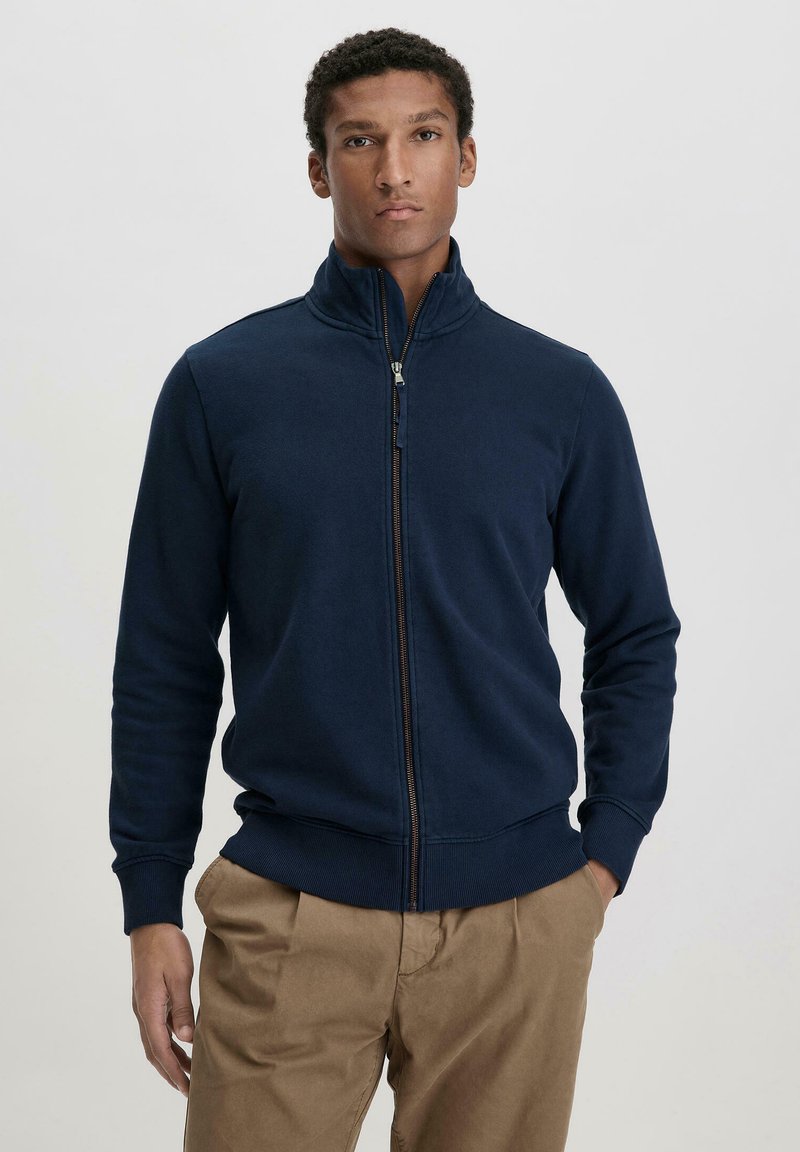 Marineblaues Zip-Up Sweatshirt aus weichem, strukturiertem Material mit hohem Kragen und gerippten Bündchen, kombiniert mit hellbraunen Hosen.