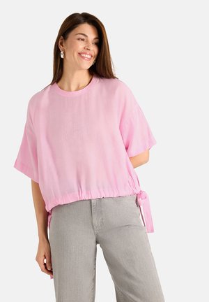 Woman smiling wearing a loose pink short-sleeve top with side ties and light gray high-waist jeans against a plain background.