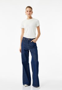Hvid kortærmet skjorte med hæklede kanter, parret med højtaljede mørkeblå flared jeans, stående på en ensfarvet hvid baggrund.