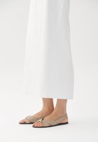 Beige lær sandaler med kryssrem-design og svart såle, med åpen tå og slingback-stil. Plassert mot en hvit bakgrunn.
