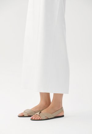 Beige lær sandaler med kryssrem-design og svart såle, med åpen tå og slingback-stil. Plassert mot en hvit bakgrunn.