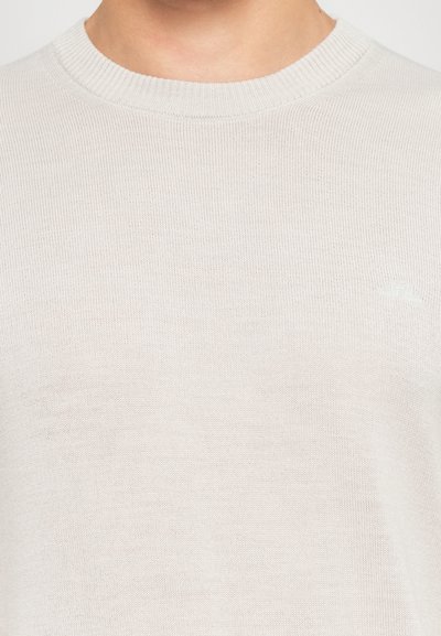 Nahaufnahme einer Person, die einen hellbeigen, gerippten Strickpullover mit rundem Halsausschnitt und einem dezenten, gestickten Logo auf der Brust trägt.