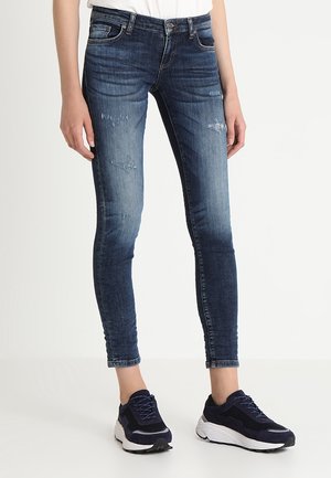 Woman wearing blue skinny distressed jeans and navy blue sneakers, standing against a plain light background.