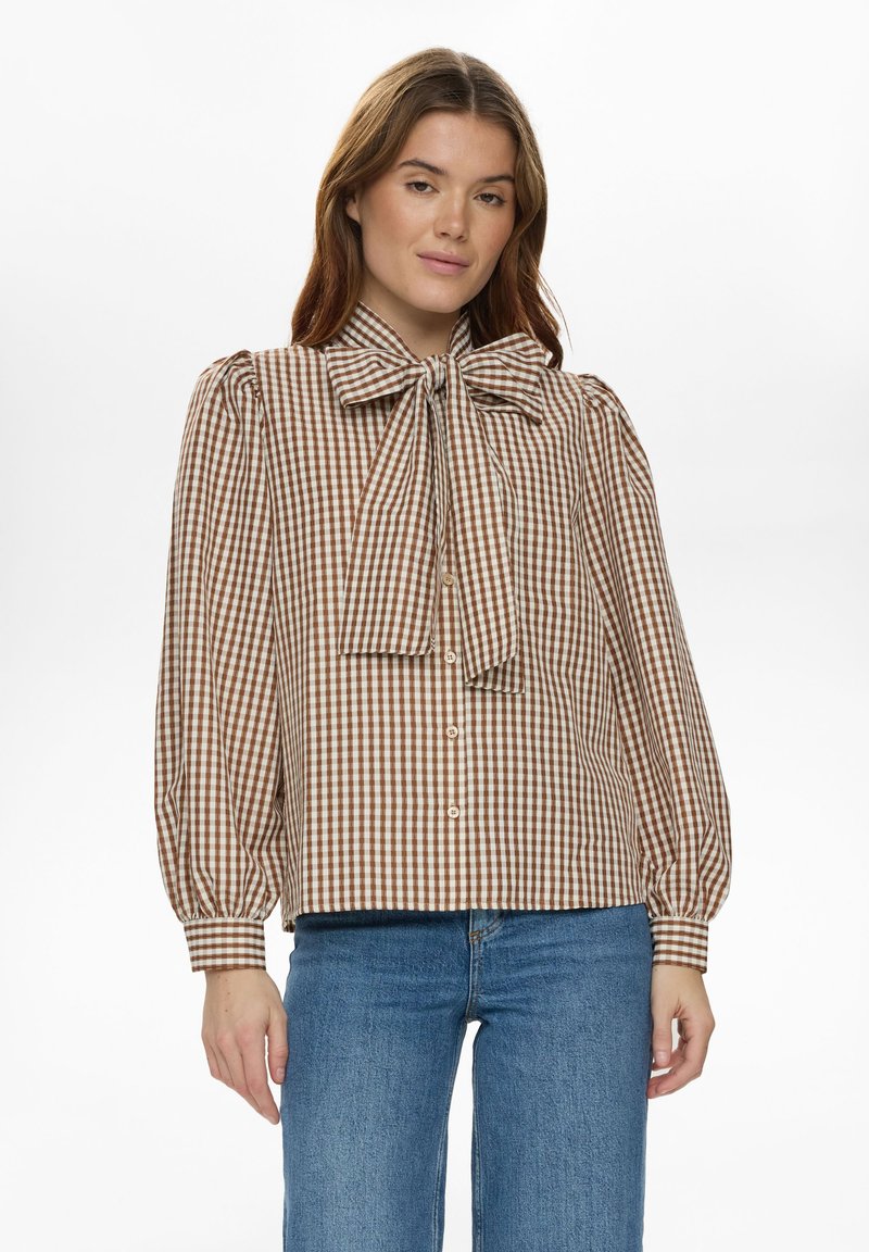 Blouse à carreaux marron et crème avec un détail de nœud papillon, manches longues bouffantes, fermeture à boutons, et tissu texturé. Associée à un jean bleu.