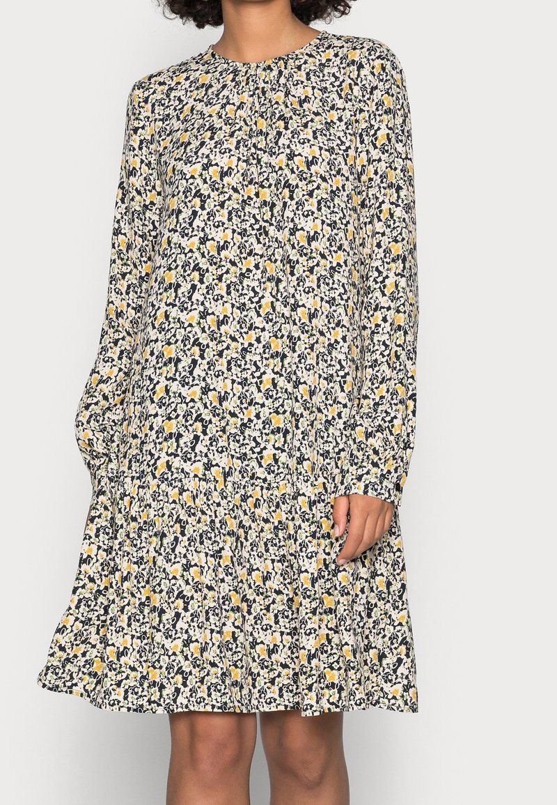 Femme portant une robe à manches longues, longueur genou, avec un motif floral dense en noir, blanc et tons jaunes sur un fond clair.