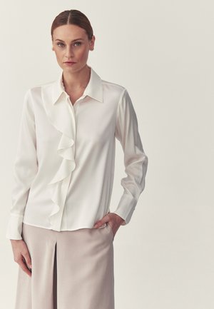 Vrouw met haar naar achteren gebonden, gekleed in een witte blouse met lange mouwen en een ruche aan de voorkant, en een beige rok, staande tegen een effen achtergrond.