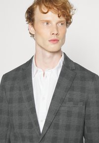 Grijze geruite blazer van textuurstof, met revers en een enkele borstzak, gedragen over een witte shirt.