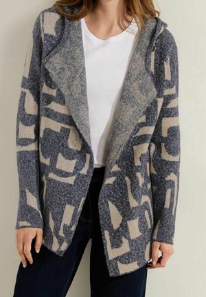 Frau trägt eine grau-beige gemusterte offene Strickjacke über einem weißen Oberteil und dunklen Hosen, steht vor einer schlichten hellen Wand.
