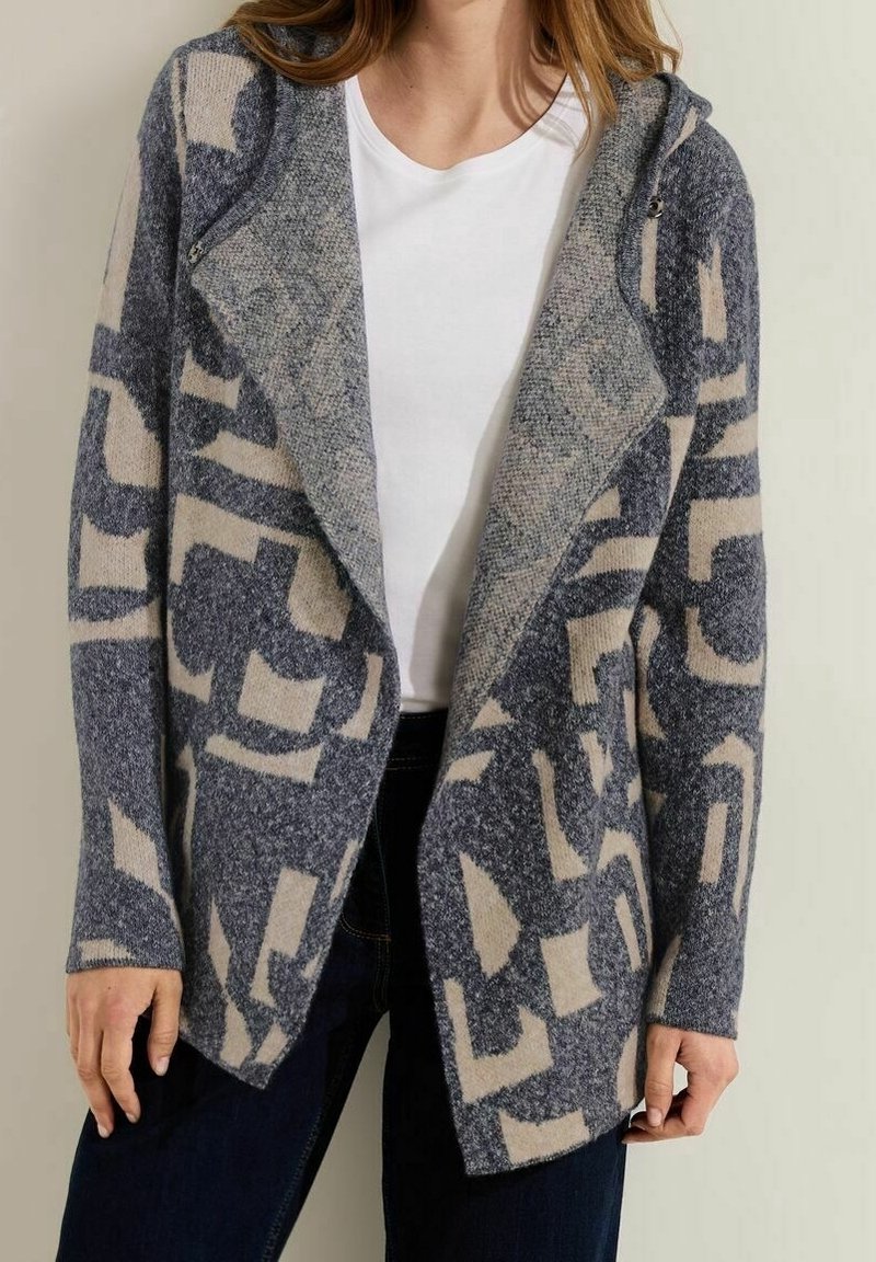 Femme portant un cardigan à motifs gris et beige ouvert sur un haut blanc et un pantalon foncé, debout contre un mur clair uni.