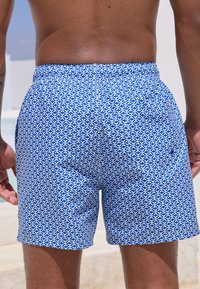 Blå badebukser med mønster og hvide geometriske designs, elastisk taljebånd og sidelommer, lavet af et let, hurtigtørrende stof.