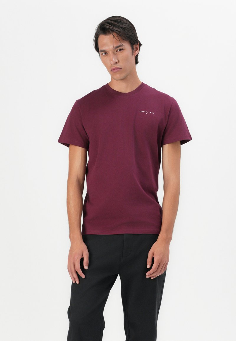 Burgunderfarbene Baumwoll-T-Shirt mit kurzen Ärmeln, Rundhalsausschnitt und kleinem weißen "Tommy Jeans" Logo auf der linken Brust. Kombiniert mit schwarzen Hosen.