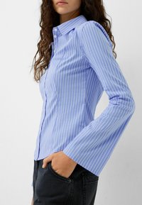 Jasnoniebieska koszula z długim rękawem w białe pionowe paski, z klasycznym kołnierzykiem i rozszerzanymi rękawami, w połączeniu z czarnymi spodniami.