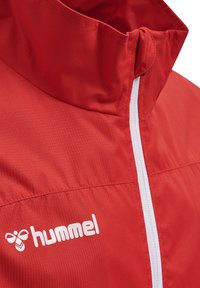 Röd sportjacka i strukturerad tyg med hög krage, vit dragkedja och logotyp i vitt. Nära vy av design detaljer.