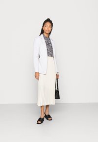 Blazer branco sobre uma camisa estampada em preto e branco, combinado com uma saia plissada creme e mules pretos de bico aberto. Segurando uma bolsa preta.