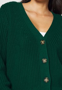 Grön stickad cardigan med V-hals, prydd med tre stora knappar och ribbad textur på ärmar och kropp.