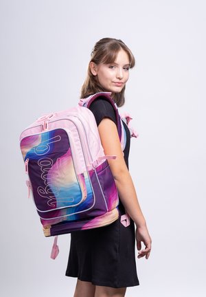Teenager-Mädchen trägt ein schwarzes Kleid und einen großen pinken und mehrfarbigen Rucksack und schaut seitlich vor einfachem Hintergrund.