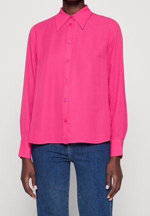 Kobieta ubrana w jaskraworóżową koszulę na guziki z długim rękawem i kołnierzykiem z ostrymi końcami oraz niebieskie dżinsy, stojąca na tle jednolitego jasnego tła.