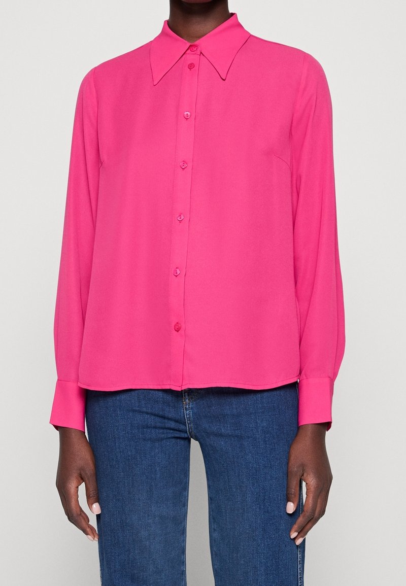 Femme portant une chemise rose vif à manches longues avec col pointu et un jean bleu, debout devant un fond clair uni.