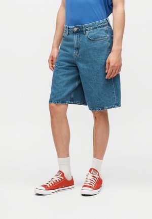 Άνδρας φορώντας μπλε τζιν σορτς μήκους μέσης, λευκές κάλτσες και κόκκινα κόκκινα χαμηλά παπούτσια, στέκεται μπροστά σε απλό φόντο.