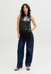 Musta paljettivaatteinen hihaton toppi, jossa on fringet, yhdistetty tummansinisiin leveälahkeisiin farkkuihin, joissa on koristeelliset hopeiset niitit, ja mustiin korokepolkimiin.