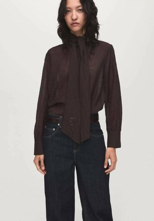 Blusa marrone semi-trasparente con maniche lunghe e collo a cravatta. Abbinata a jeans in denim scuro con cintura e vestibilità rilassata.