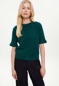 Maglione corto a maniche corte in maglia verde, scollatura rotonda, texture morbida, vestibilità rilassata e orlo a coste. Abbinato a pantaloni neri.