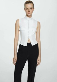 Top blanco sin mangas con cuello alto, cierre frontal y dos bolsillos en el pecho, combinado con pantalones negros de corte ajustado, que presentan una silueta ceñida.
