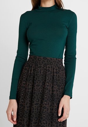 Woman wearing a fitted dark green long-sleeve turtleneck top paired with a high-waisted, black leopard print skirt.