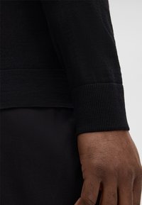 Zwarte gebreide sweater mouw met ribbels aan de boord, met een gladde textuur en een aansluitend ontwerp. Hand zichtbaar tegen een donkere achtergrond.