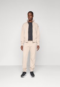 Femme portant un survêtement beige avec un blouson zippé, des baskets noires et un T-shirt noir, se tenant devant un fond gris clair uni.