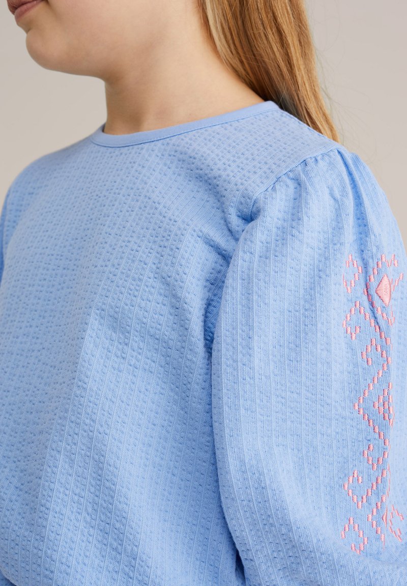 Enfant portant un haut bleu clair texturé avec manches bouffantes et broderie géométrique rose sur le haut du bras, de profil.