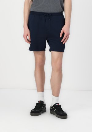 Άνδρας που φοράει ναυτική μπλε βερμούδα με κορδόνια, γκρι μπλουζάκι, άσπρες κάλτσες τύπου crew και μαύρα αθλητικά παπούτσια, στέκεται μπροστά σε λευκό φόντο.