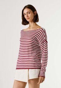 Junge Frau, die einen rot-weiß gestreiften Langarmpullover und weiße Shorts trägt, vor einem schlichten hellen Hintergrund stehend.