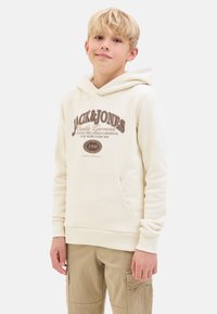Sweatshirt crème avec une poche avant, présentant un texte de logo marron. Manches longues, tissu doux et capuche avec cordon. Associé à un pantalon cargo kaki.